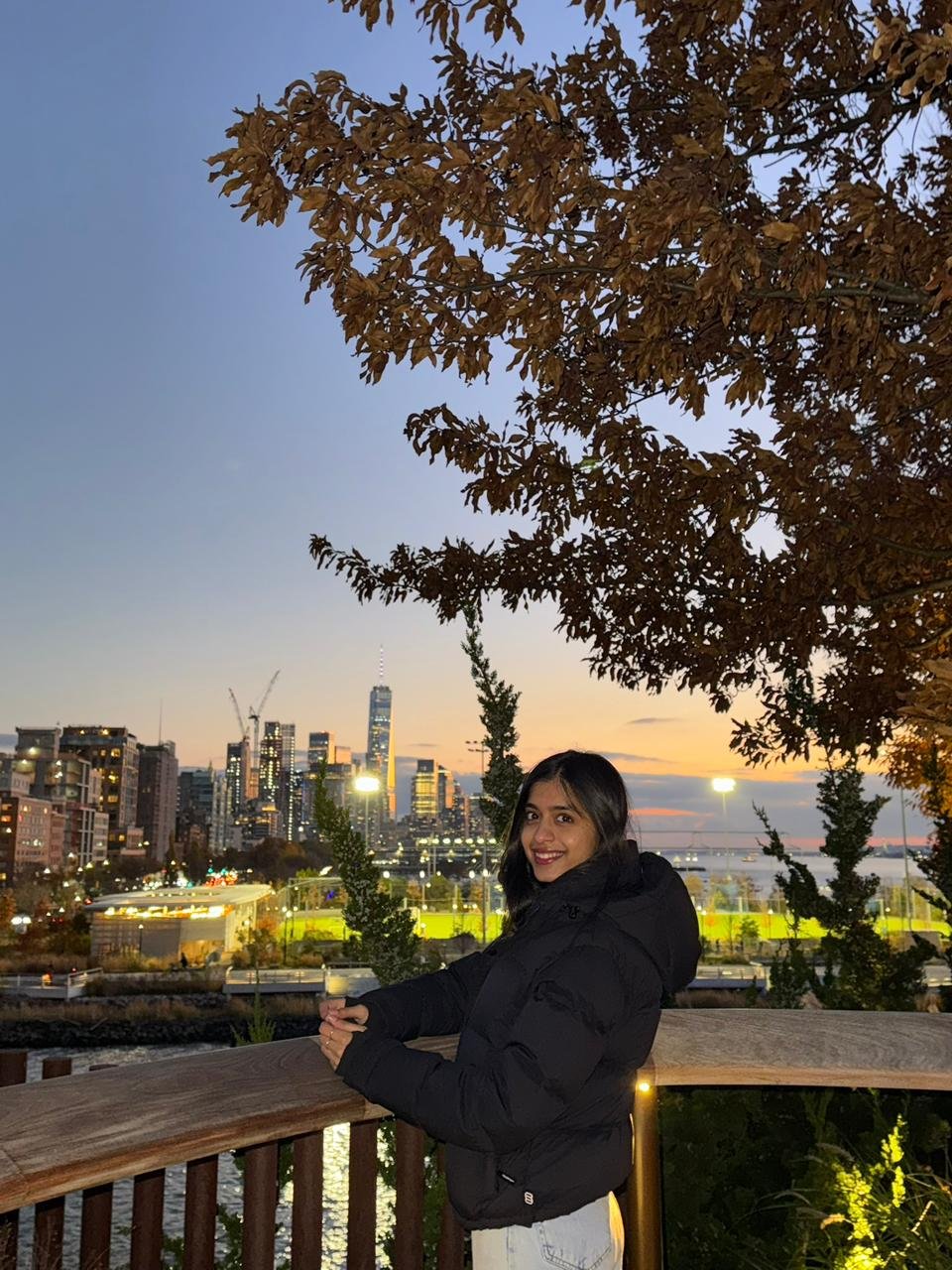 Ananya Rajora smiling on a waterfront boardwalk at dusk with a city skyline in the background